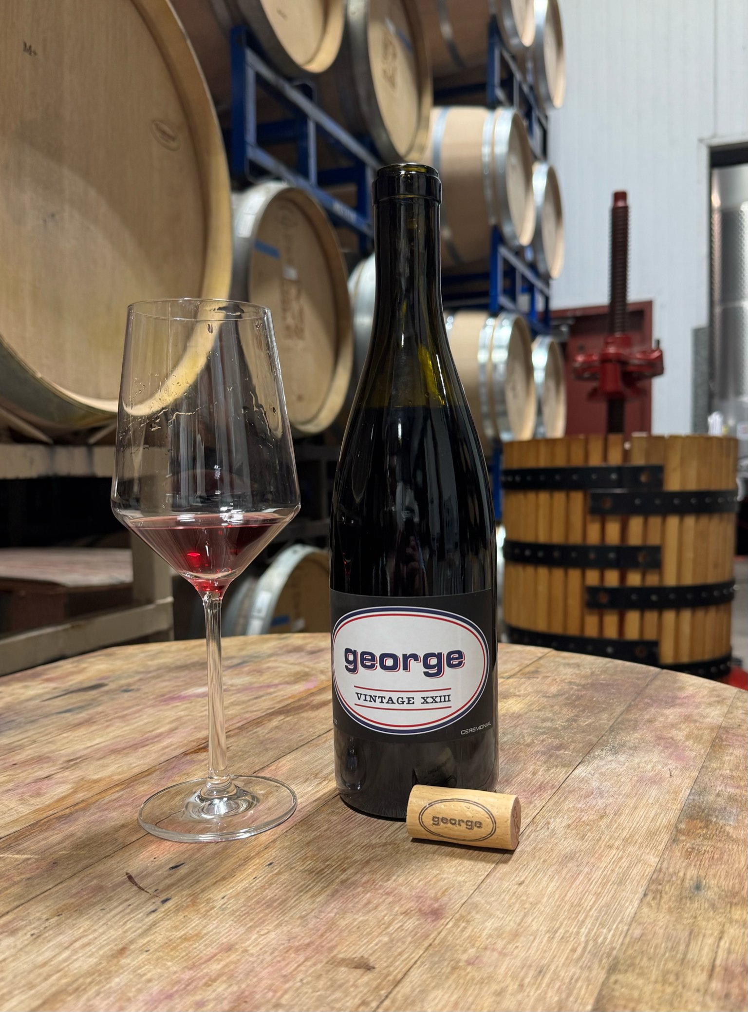 A bottle of George Pinot Noir beside a poured glass and a George cork on a wooden table in the barrel room
